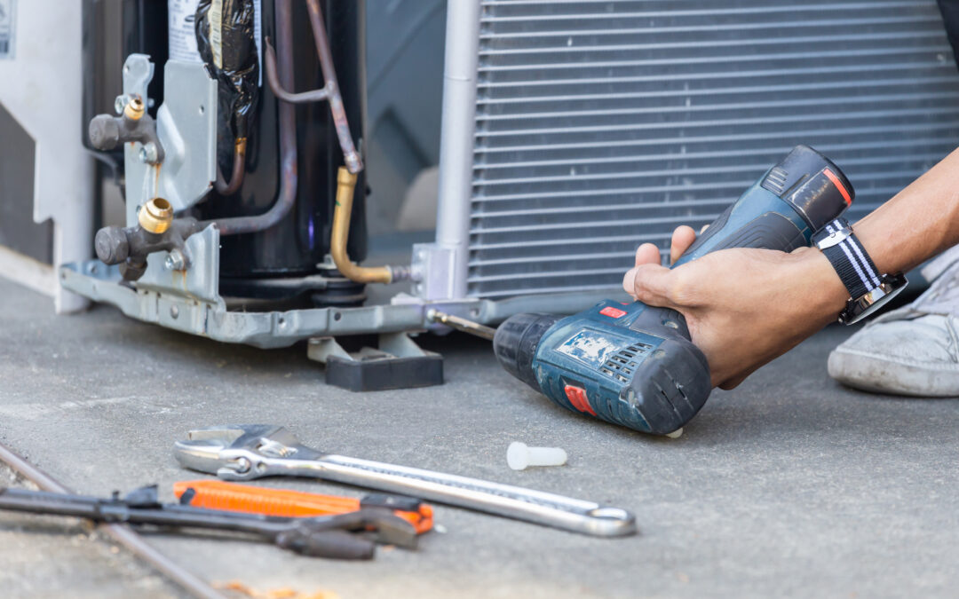 Hand using a power drill to repair an air conditioning unit, with tools like pliers and wrenches visible on the ground, illustrating AC repair services in Sacramento.